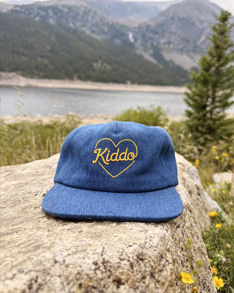A blue denim-style flat-brim cap with a yellow embroidered heart and the word 'Kiddo' inside, resting on a large rock with a mountain lake landscape in the background.