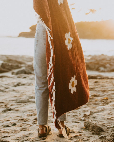 A person wearing a Lazy Daisy Blanket on the beach, showcasing its vibrant design and midweight fabric.
