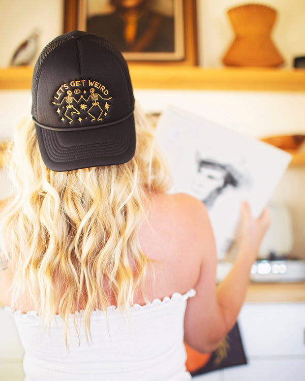 A woman wearing a black foam trucker hat with the phrase Let's Get Weird, showcasing a fun and unique style.