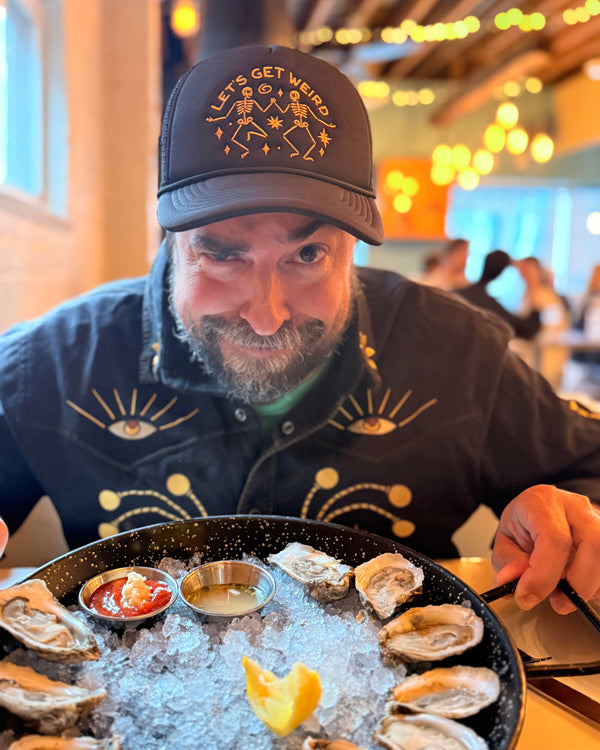 Man wearing a Let's Get Weird Hat, enjoying oysters at a vibrant restaurant, showcasing unique style.