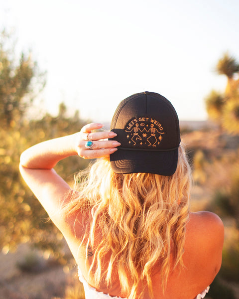Woman wearing a Let's Get Weird Hat, showcasing its unique design against a sunset background.