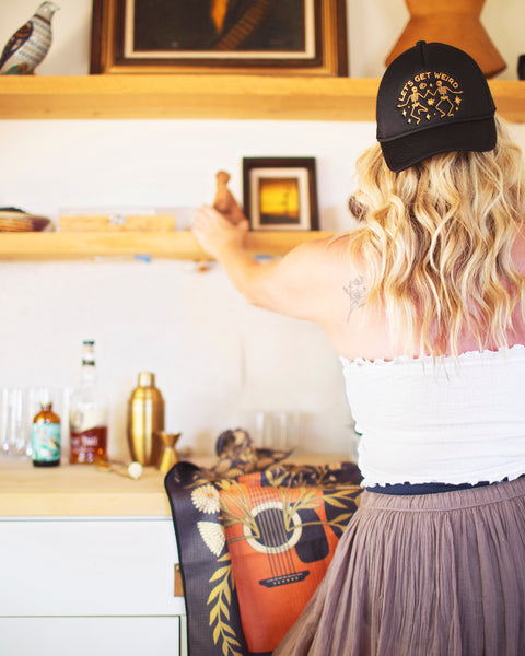 Person wearing a Let's Get Weird Hat while arranging items on a shelf in a cozy setting.