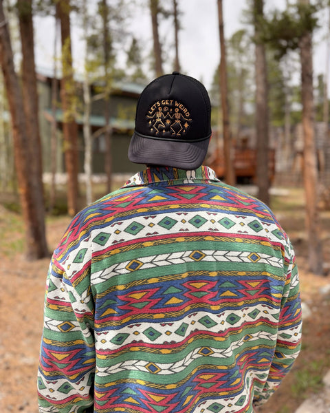 Person wearing a colorful shirt and a Let's Get Weird Hat in a forest setting.