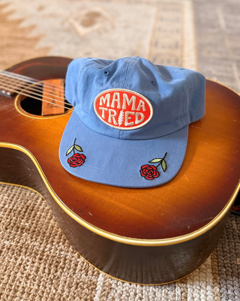 Mama Tried Hat displayed on a guitar, featuring vibrant red roses and an eye-catching logo.