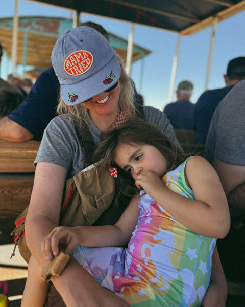 Woman wearing a Mama Tried Hat while holding a child in a colorful dress, enjoying a casual outdoor setting.