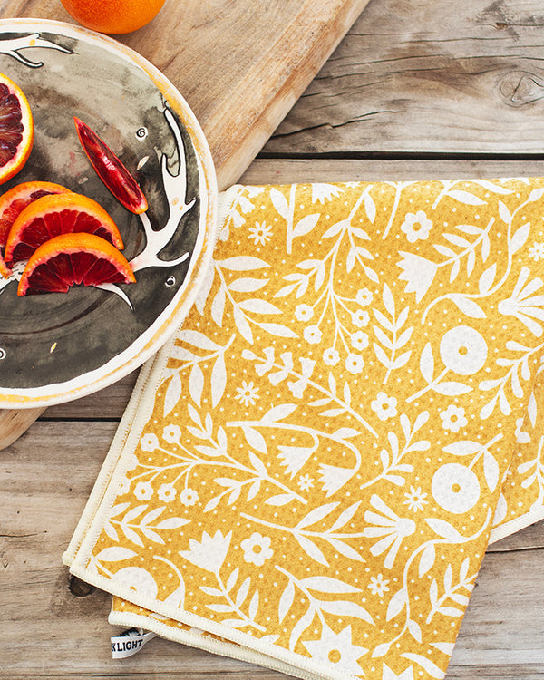 Yellow Meadow Kitchen & Tea Towel made from recycled materials, featuring a floral design on a wooden table next to a plate of citrus fruits.