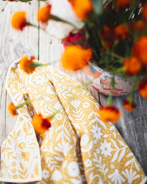 Meadow Kitchen & Tea Towel draped on a wooden surface with vibrant orange flowers in the background.