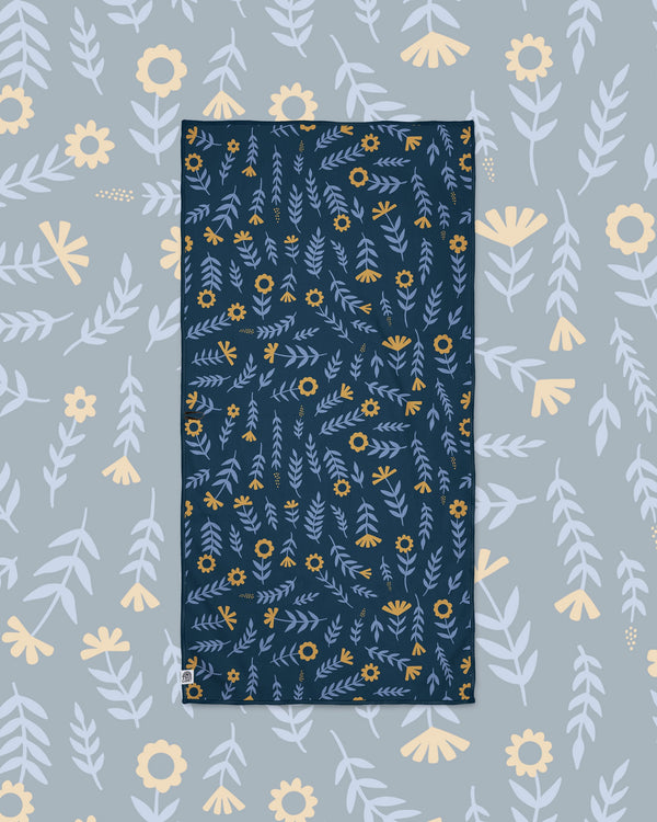 Mermaids Wander & Travel Towel in navy with blue leaves and gold flowers, laid flat on a gray-blue floral background.
