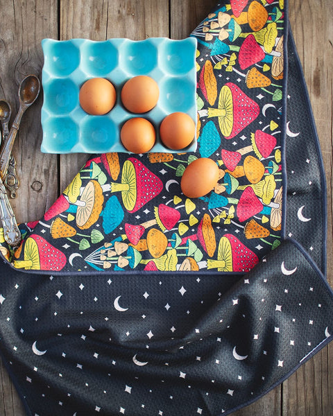 Midnight Mushroom Kitchen & Tea Towel with vibrant mushroom design, beside eggs in a blue tray and vintage spoons on a wooden table.