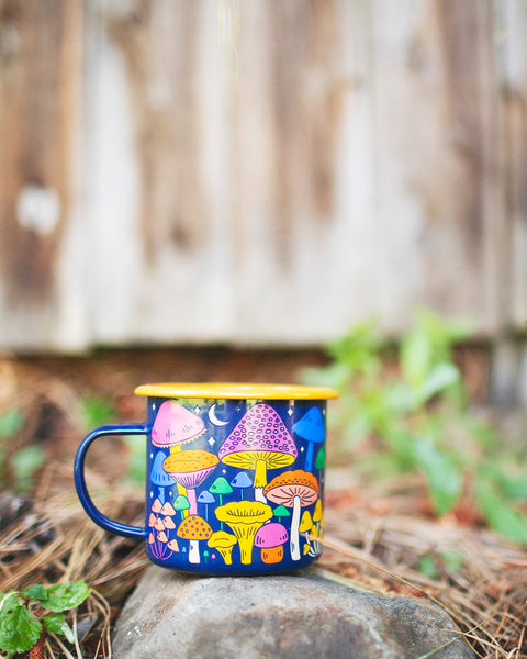 Midnight Mushroom Enamel Mug 16oz on a rock with a chicken in the background, showcasing its vibrant mushroom design.