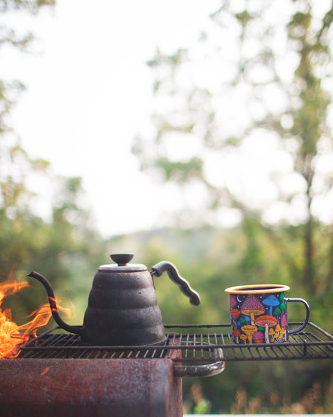 Midnight Mushroom Enamel Mug 16oz next to a kettle on a grill outdoors with trees in the background.