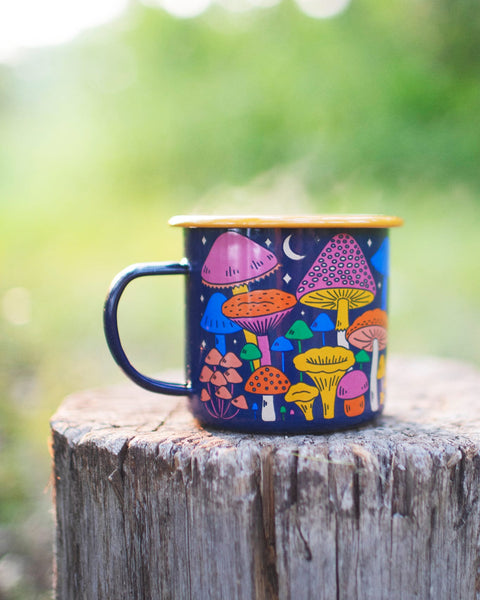 Midnight Mushroom Enamel Mug 16oz with colorful fungi design, resting on a wooden stump. Trek Light, perfect for outdoor adventures.
