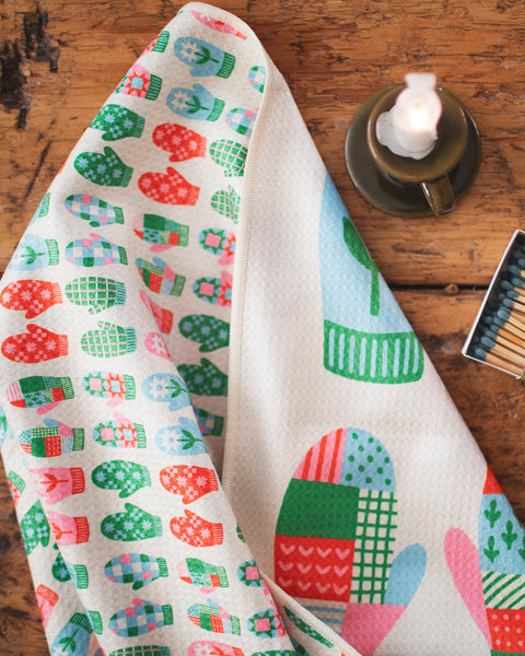 Mittens Kitchen & Tea Towel featuring colorful mitten designs on a wood table with a candle and matchbox.