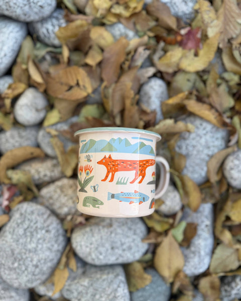 Mountain Stream Enamel Mug 16oz on a bed of rocks and autumn leaves, featuring vibrant animal and nature designs.
