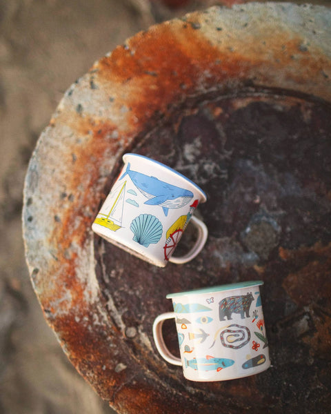 Mountain Stream Enamel Mug 16oz displayed on a rustic surface with colorful ocean-themed designs.
