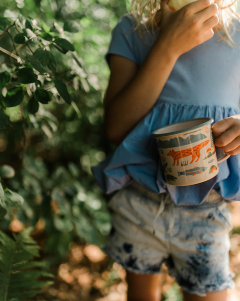 A person holding a Mountain Stream Enamel Mug 16oz among lush greenery, perfect for outdoor sipping.