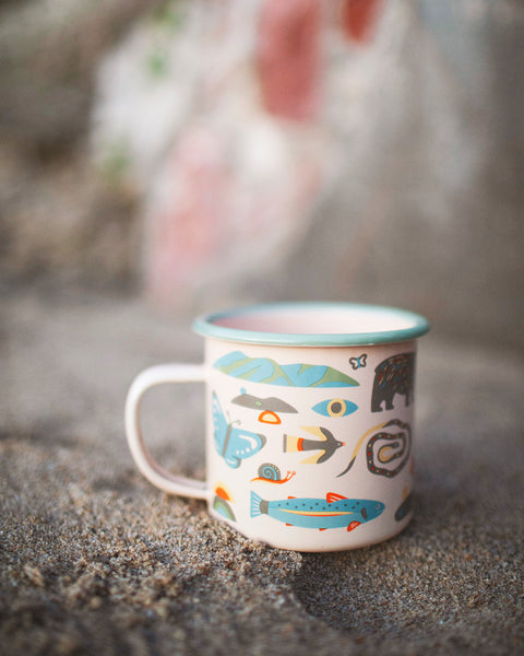 Mountain Stream Enamel Mug 16oz with colorful nature-inspired design on sandy surface.