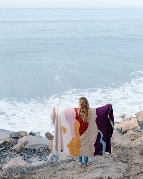 A woman holding the Mountain Sunrise Blanket by the ocean, showcasing the colorful design against a tranquil sea.