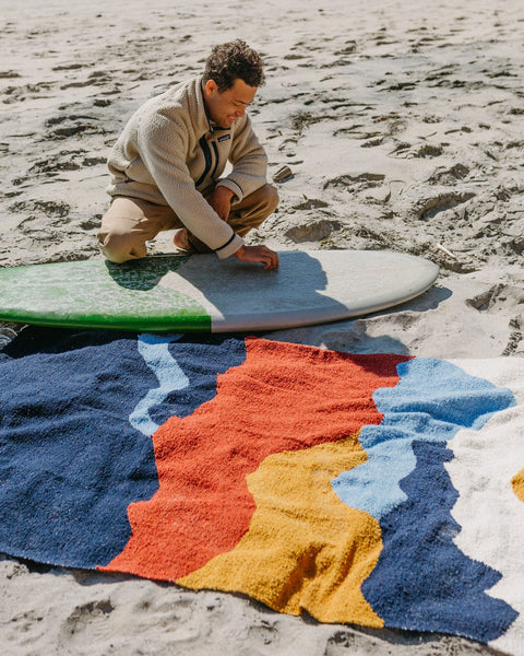 Man on beach admiring colorful Trek Light Mountains Blanket near surfboard, handwoven from recycled materials. Cozy and eco-friendly.