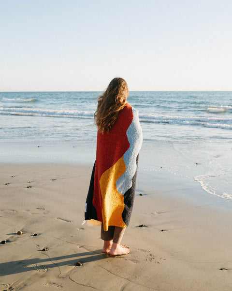 Woman wrapped in colorful Trek Light Mountains Blanket standing on sandy beach facing ocean waves.