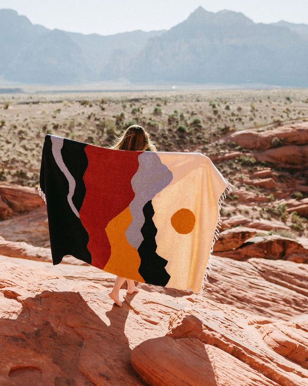 Person holding a colorful handwoven Mountains Blanket by Trek Light, set against a stunning mountain backdrop, made from recycled materials.