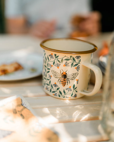Honey Bee Enamel Mug 16oz resting on a table surrounded by pastries and soft morning light.