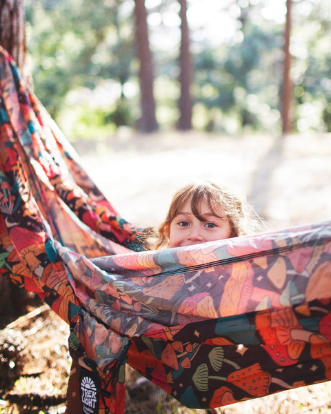 A child peeking out from a colorful Double Hammock & Pro Hanging Straps (Midnight Mushroom Print) in a forest.
