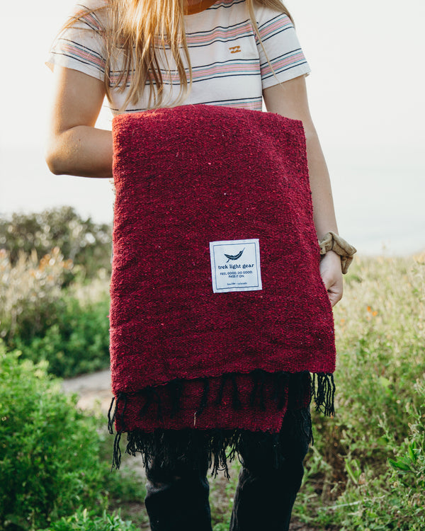 Person holding a red Trek Light Napa Blanket outdoors, made from recycled materials. Eco-friendly and cozy for naps and wine nights.