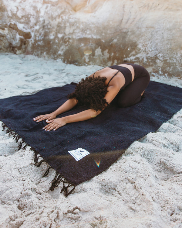 Person practicing yoga on navy Newport Blanket by Trek Light Gear, set on sandy beach.