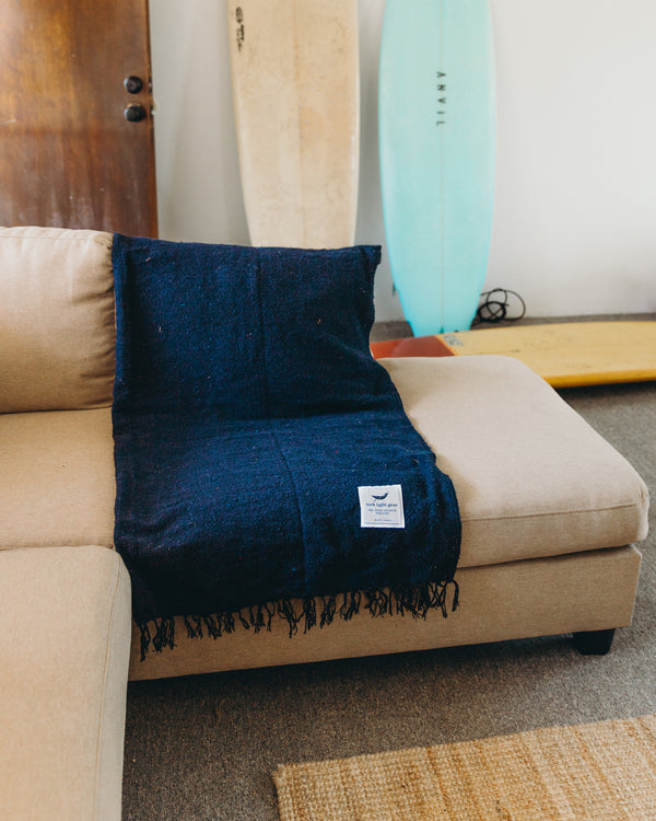 Navy Newport Blanket from Trek Light draped over a beige couch, with surfboards in the background, embodies coastal comfort.