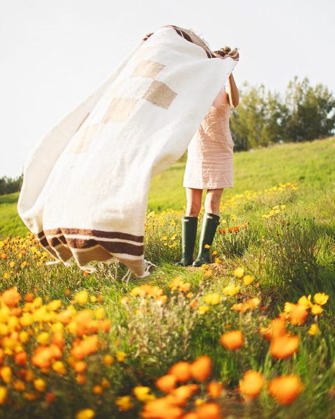 Person joyfully holding a cozy, handwoven North Star Blanket made from recycled clothing materials in a vibrant flower field. By Trek Light.