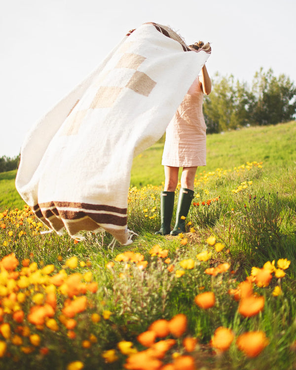 Person joyfully holding a cozy, handwoven North Star Blanket made from recycled clothing materials in a vibrant flower field. By Trek Light.