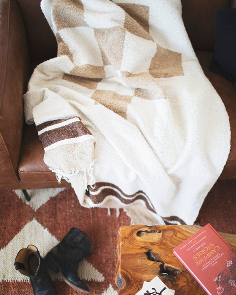 The handwoven North Star Blanket in cream and tan draped over a leather sofa above a rug and boots, Trek Light