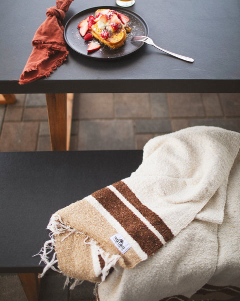The handwoven North Star Blanket in cream and brown folded over a patio bench beside a breakfast plate, Trek Light