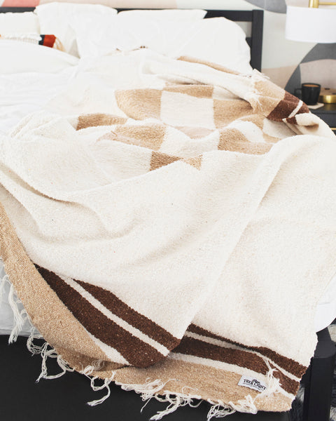 The handwoven North Star Blanket in cream and tan draped across a made bed with white bedding, Trek Light