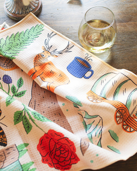 Oregon Kitchen & Tea Towel featuring vibrant designs with a deer and coffee cup on a rustic table.