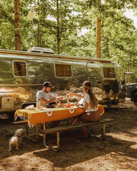 Couple enjoying a picnic on a Peace Blanket outside their Airstream in nature, surrounded by trees.