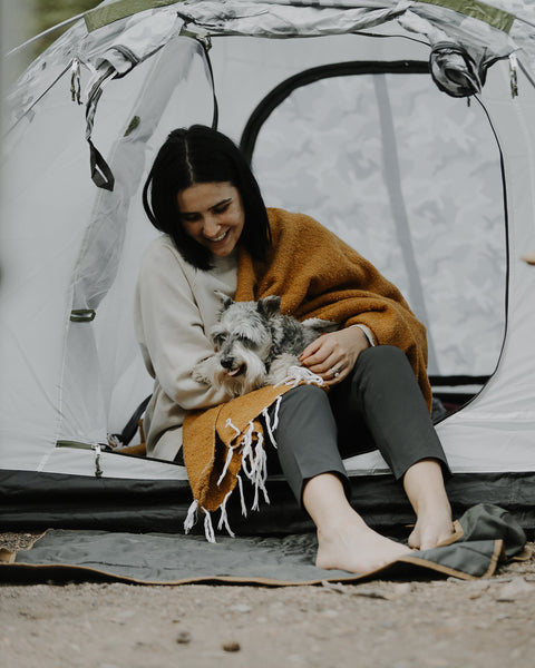 Woman sitting in a tent with her dog, wrapped in a cozy Peace Blanket. Perfect for camping and comfort.