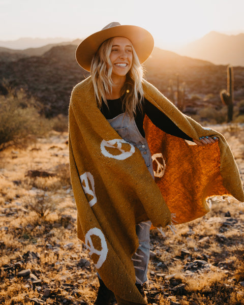 Woman in a wide brim hat wrapped in the handwoven gold Peace Blanket from Trek Light in the desert at sunset