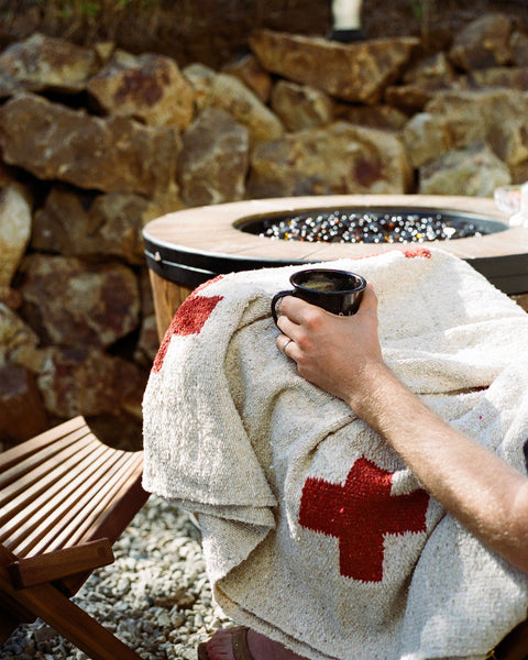 Person holding a cup while wrapped in a Positive Vibrations Blanket next to a fire pit.