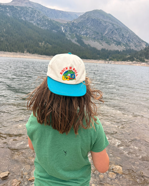 Child wearing a Rock N Roll Kids Hat while fishing by a lake, surrounded by mountains and nature.