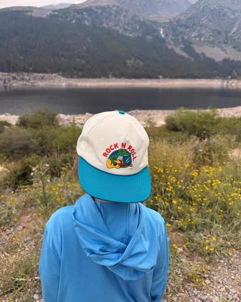 Child wearing a Rock N Roll Kids Hat while enjoying nature by a lake and mountains.