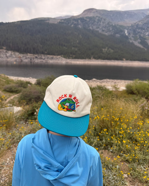 Child wearing a Rock N Roll Kids Hat in a scenic outdoor setting with mountains and flowers.