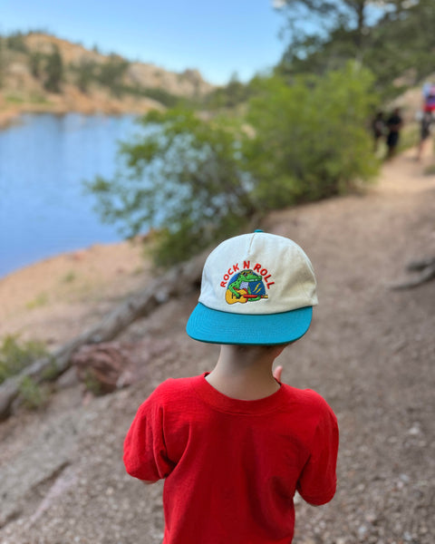 Child wearing a Rock N Roll Kids Hat by the water, capturing memories on an adventure.