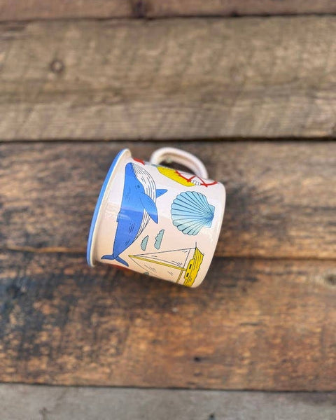 Salty Seas Enamel Mug 16oz featuring nautical designs with whales, seashells, and sailboats on wooden background.