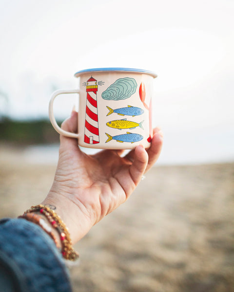 Hand holding a Salty Seas Enamel Mug 16oz with nautical lighthouse and fish design, perfect for camping and cozy mornings.