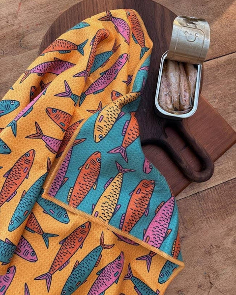 Sardine School Kitchen & Tea Towel on a wooden board with a tin of sardines, showcasing vibrant fish patterns.