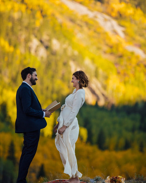 Couple exchanging vows in a vibrant autumn mountain setting, with a bride in a white dress and groom in a suit, surrounded by colorful foliage.