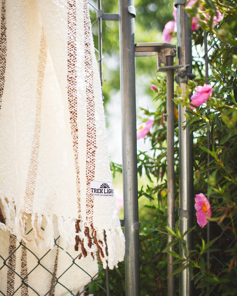 Handwoven Silverton blanket with stripes, made from recycled materials, hanging near pink flowers outdoors.