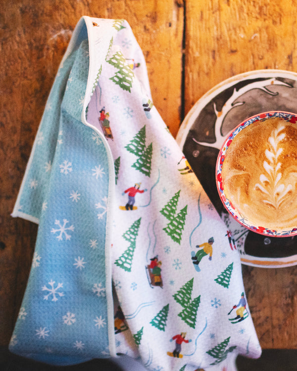 Snow Day Kitchen & Tea Towel with winter design beside a latte on a wooden table. Snowflake design, super absorbent, quick-drying kitchen towel.